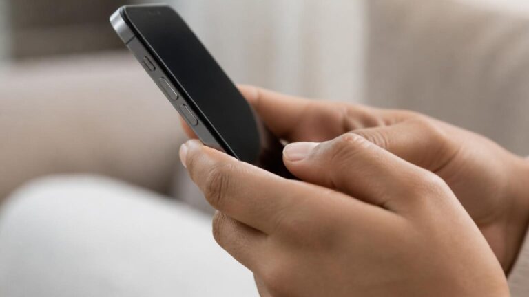 Mãos segurando um smartphone com foco nas bordas e botões laterais, em fundo neutro.